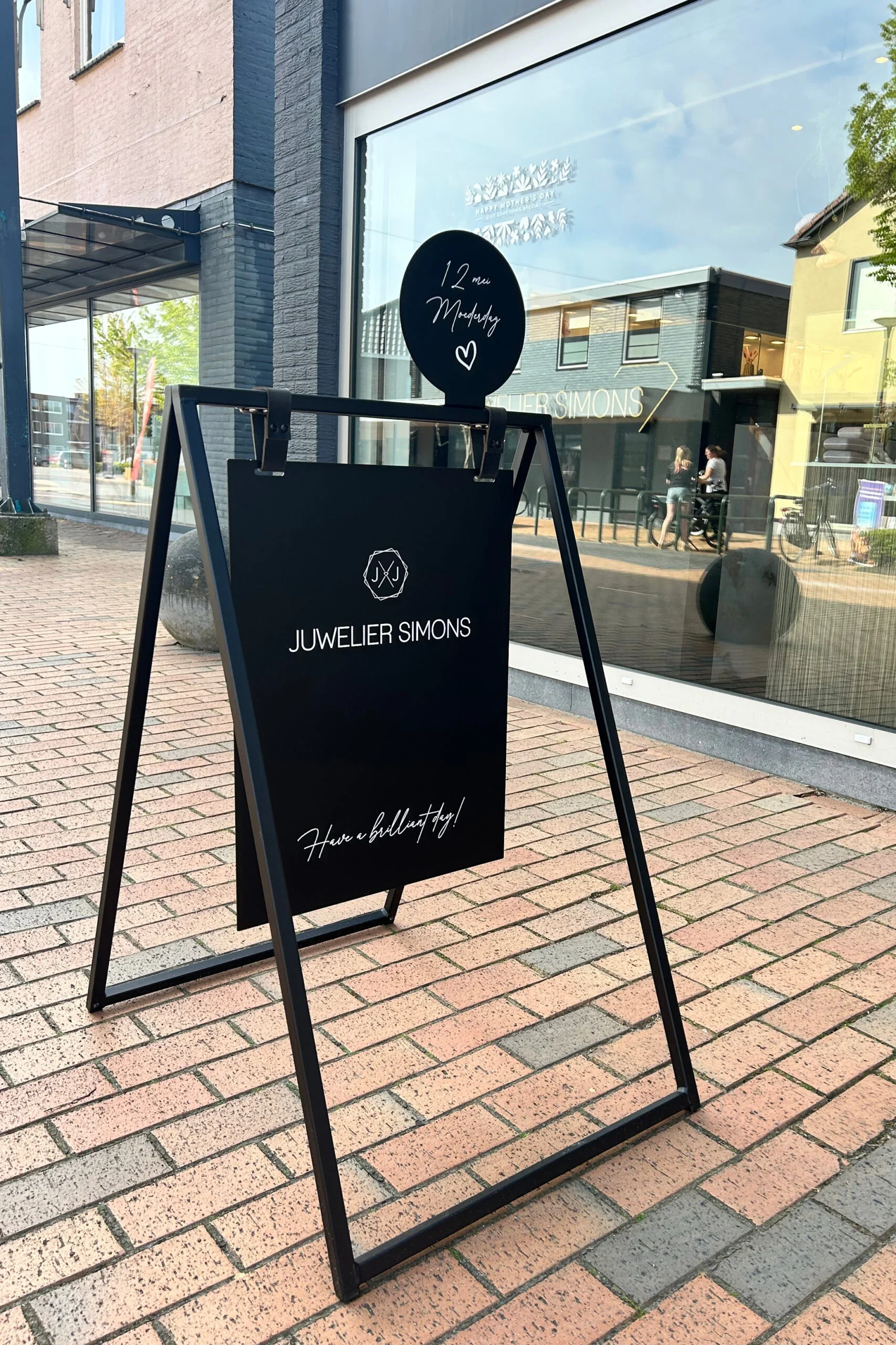 Svart reklamställ utanför en butik med texten "JUWELIER SIMONS" på en stor platta. Ovanför finns en mindre rund skylt som visar "12 mi Måndag" och ett hjärta. Mönstrade tegelplattor på golvet och en reflektion av omgivningen i butiksfönstret, där personer går förbi.