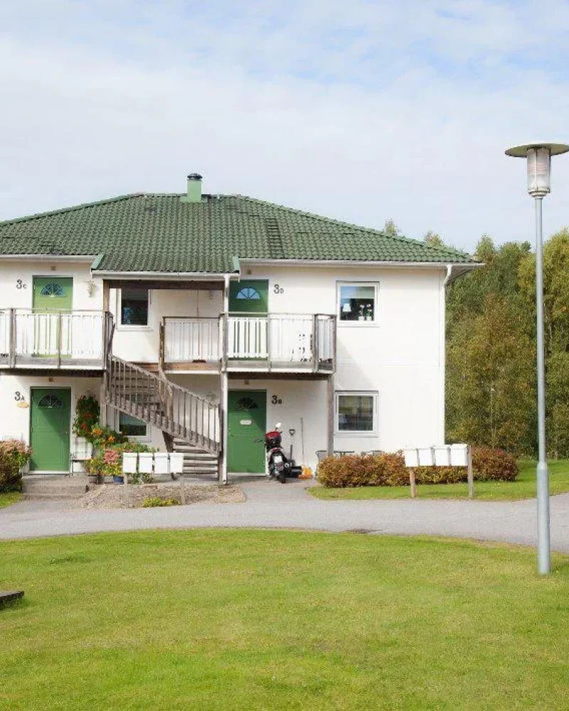 Tvåvåningshus med vit fasad och grönt tak, omgiven av grön gräsyta och buskar. Ingångar till lägenheter är på framsidan, med räcken och en trappa i mitten. I bakgrunden syns fler byggnader och träd. En gatlykta står på gräsmattan.