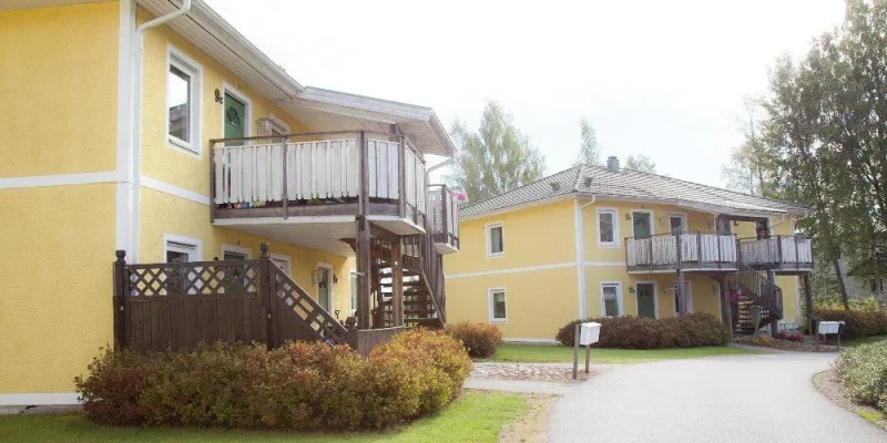 Två gula flerfamiljshus med träveranda, omgivna av grönska. Den ena byggnaden har en trappa och en pergola i mörkt trä. Stigen mellan husen är grusad och det finns buskar längs kanterna. Träden i bakgrunden ger en lantlig atmosfär, med en ljus himmel som syns ovanför.