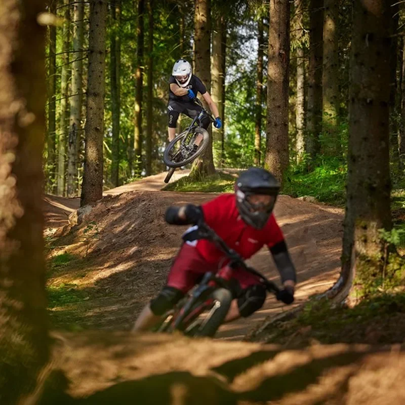 Två cyklister i skogen utför tricks på mountainbikes. Den ena cyklisten, iklädd svart tröja och hjälm, hoppar över en upphöjning medan den andra, i röd tröja och svart hjälm, svänger nerför en kurva. Runt dem finns höga träd och mjuka, grusiga stigar.