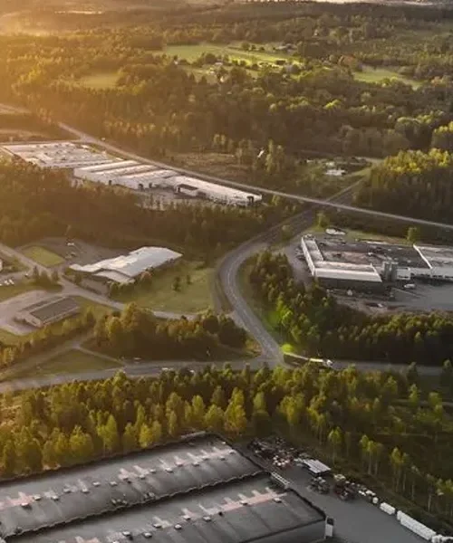 Vy över ett industriområde omgivet av grönskande landskap. I förgrunden syns lagerbyggnader med tak i grått, medan en svängd väg leder genom området. I bakgrunden sträcker sig stora fabriksbyggnader och ett fält med träd under en solig himmel, vilket ger en varm och ljus stämning.